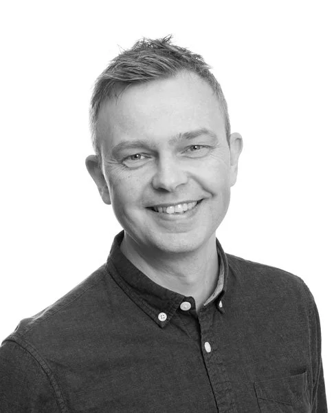 Portrettbilde av en kledd mann med kort hår som smiler, tatt i svart-hvitt.