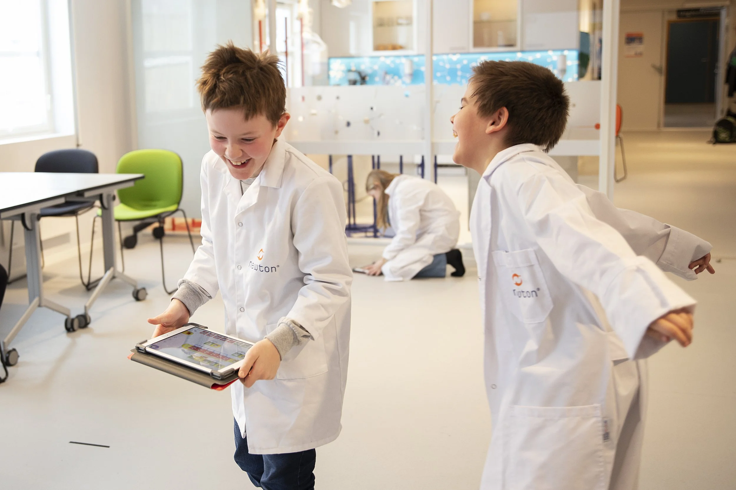 To unge gutter iført hvite laboratoriefrakker smiler og ser på en nettbrett, mens en annen gutt i bakgrunnen også er i ferd med å jobbe med noe på gulvet. De er i et Newton-rom.