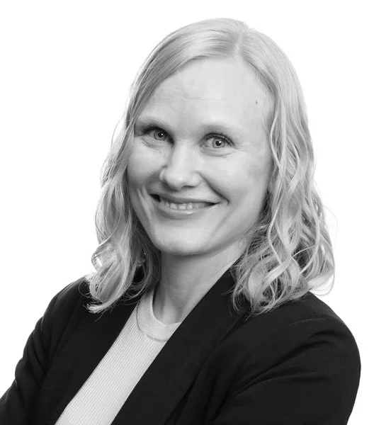 Portrett av en smilende kvinne med lys hår, iført svart jakke og lyse genser, mot en hvit bakgrunn.