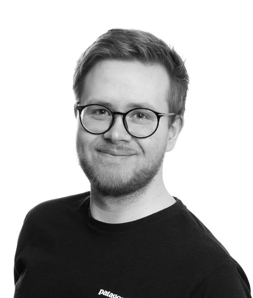 Portrett av en smilende mann med briller og kort, lyst hår, iført svart t-skjorte, i svart-hvitt fotografi.