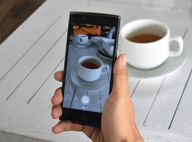 A person taking a photo of a white cup filled with tea on a white wooden table with a smartphone, with another similar cup in the background.