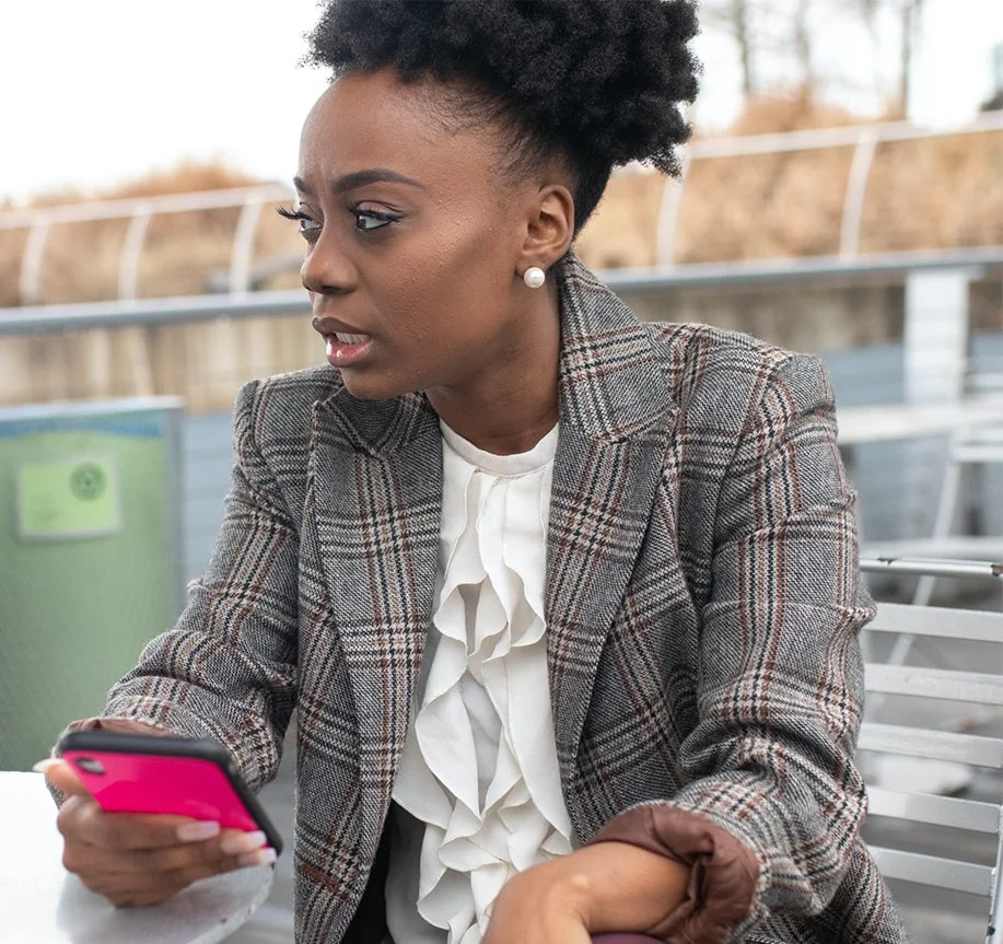 A woman sitting outdoors on a bench, looking at her pink smartphone with a concerned or focused expression. She has short, curly black hair, wears pearl earrings, a plaid blazer, and a white ruffled blouse.