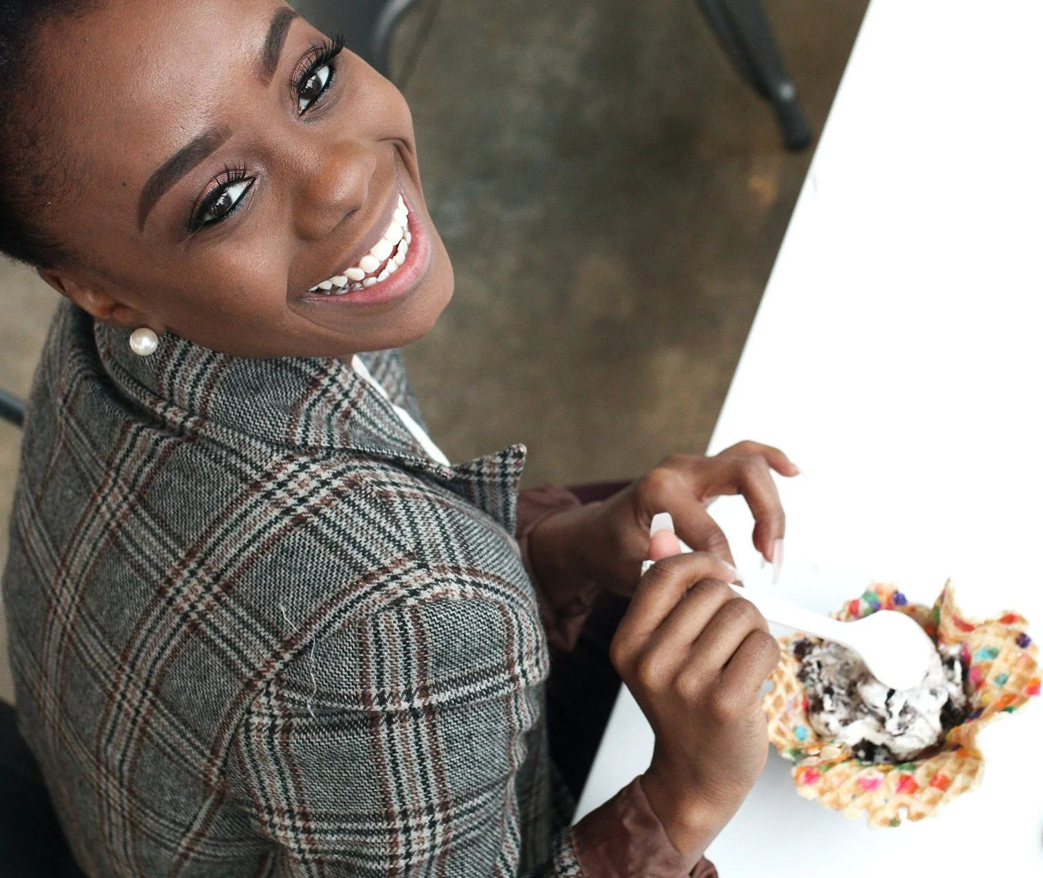 A woman with dark skin, short black hair, and pearl earrings, smiling and looking up at the camera while holding a spoon and eating ice cream from a waffle bowl with colorful sprinkles.