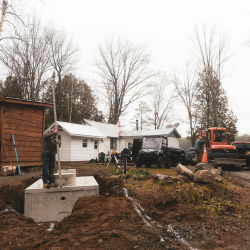 Construction workers installing a concrete foundation outside in a residential area with leafless trees, parked cars, and houses in the background.