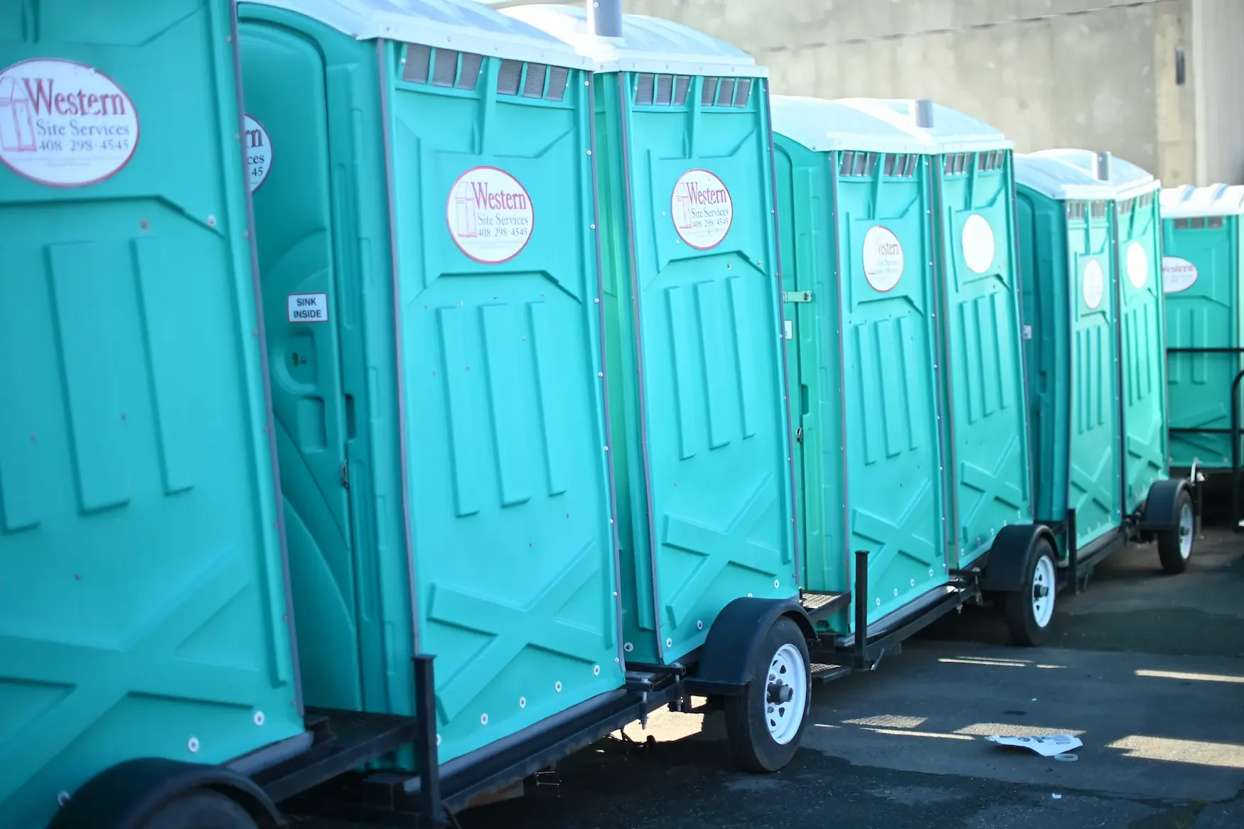 A row of portable toilets in teal color lined up outdoors on a paved surface.