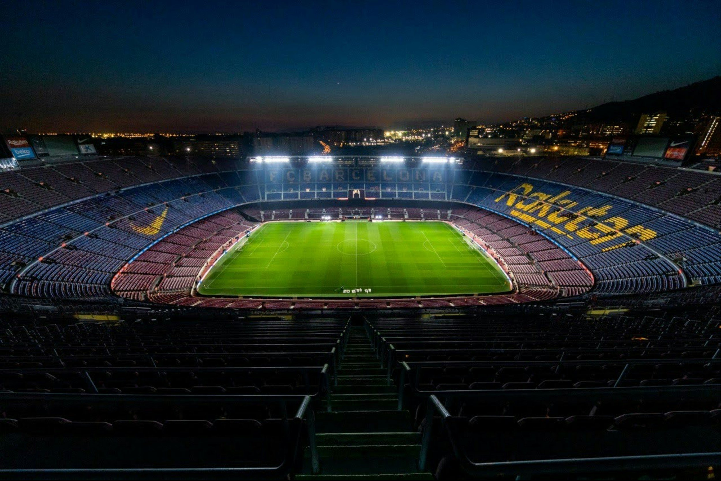 Empty soccer stadium with lights on at night, green field, and city lights in the background.