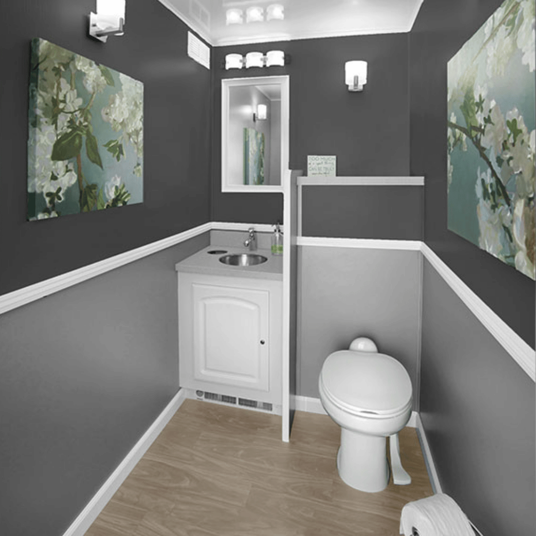 A small bathroom with dark gray upper walls and light gray lower walls, white trim, wood-look flooring, a small white vanity with a round sink, a white toilet, a mirror above the sink, two wall-mounted lights, and two floral paintings on the walls.