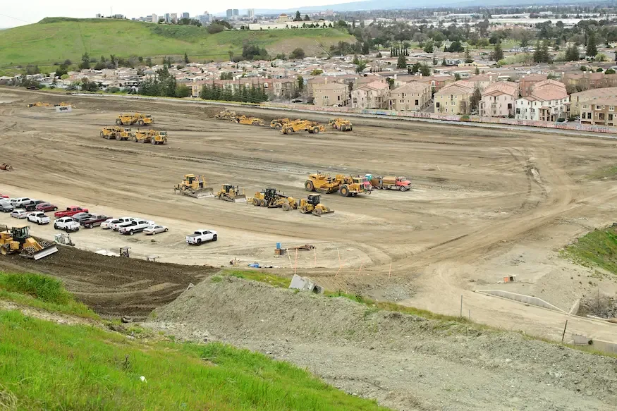 A construction site with large bulldozers and dump trucks, with parked cars at the front, and residential neighborhood homes with hills and cityscape in the background.
