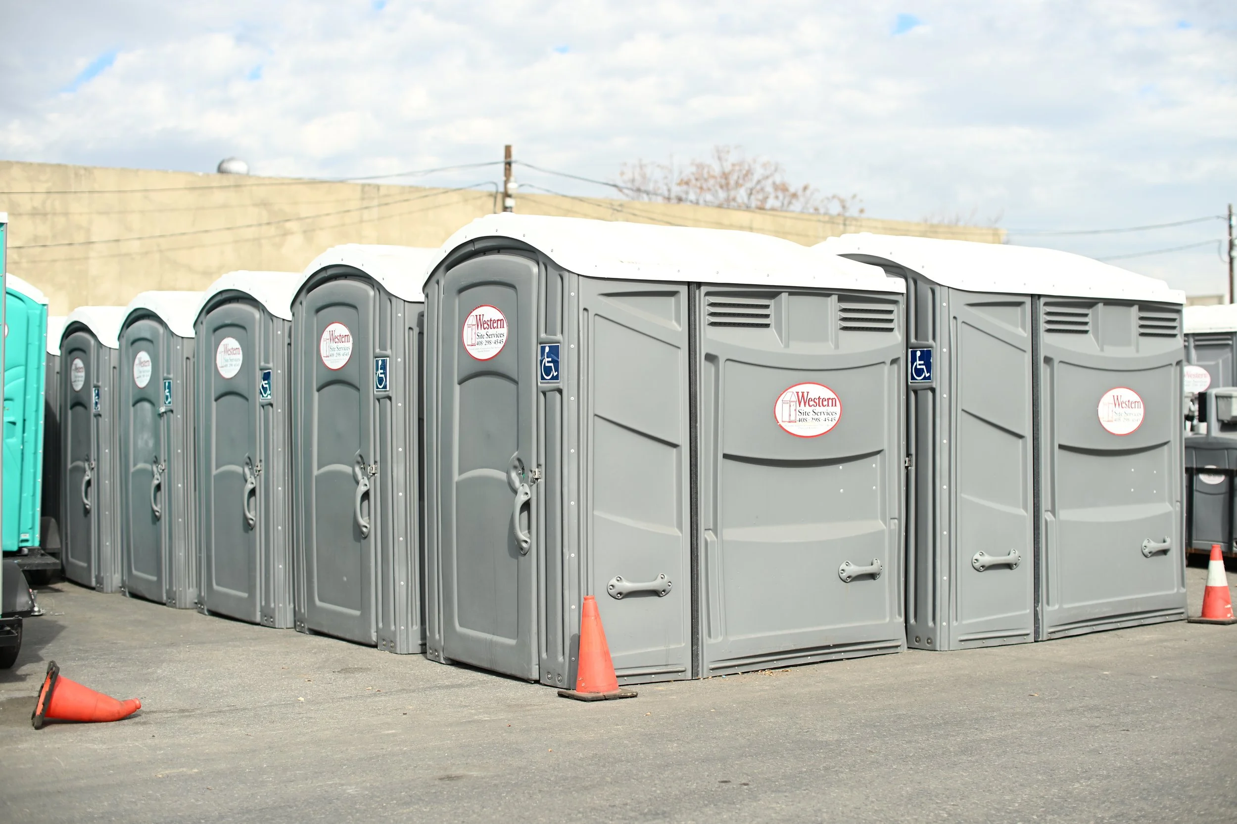 A row of portable toilets with grey doors and a handicap-accessible sign on each, set on a paved outdoor surface. Orange cones are placed nearby, and a cloudy sky is visible in the background.