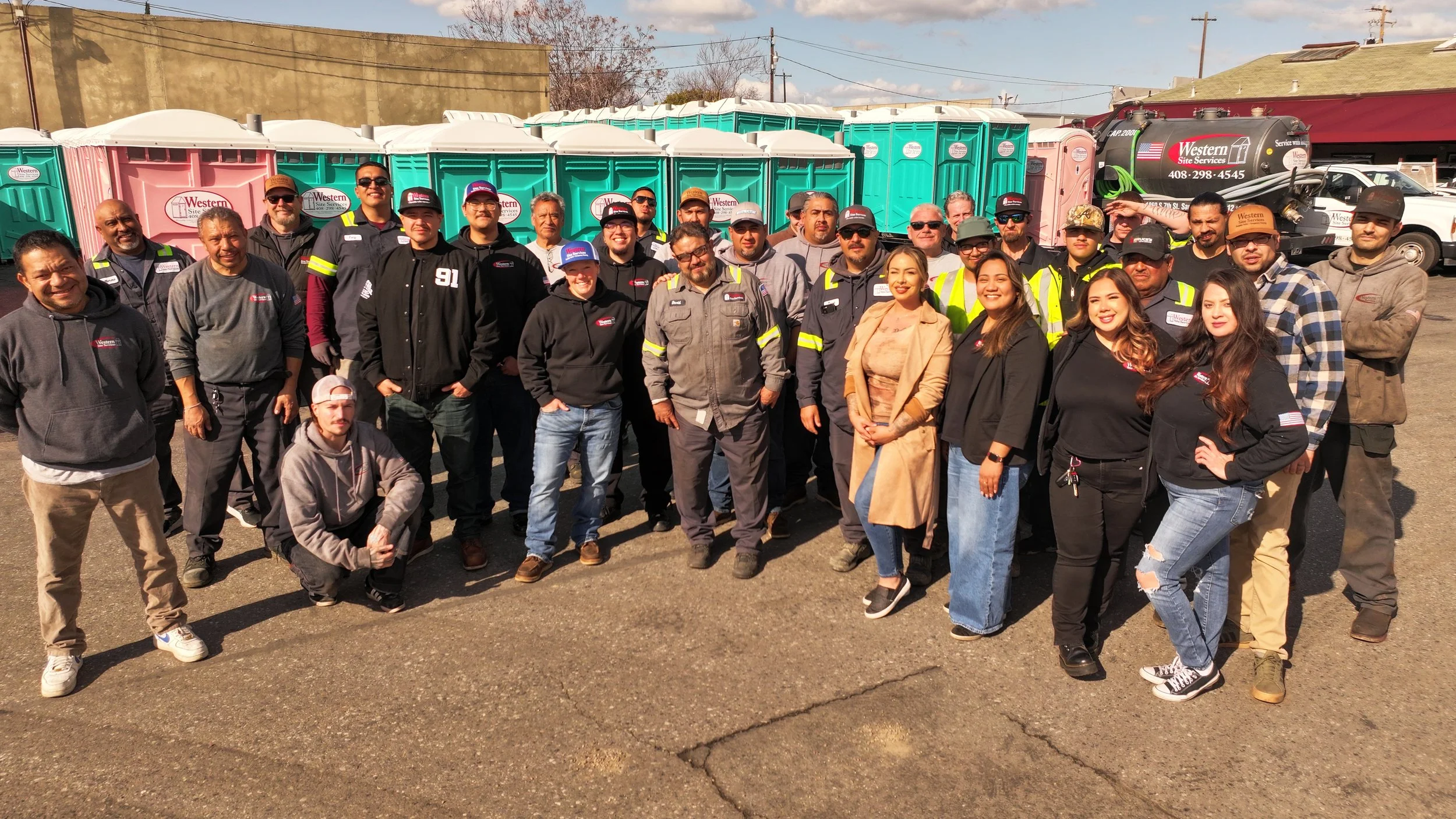 Group of people standing outdoors in front of colorful portable toilets, with some wearing company uniforms and safety gear, under a partly cloudy sky.