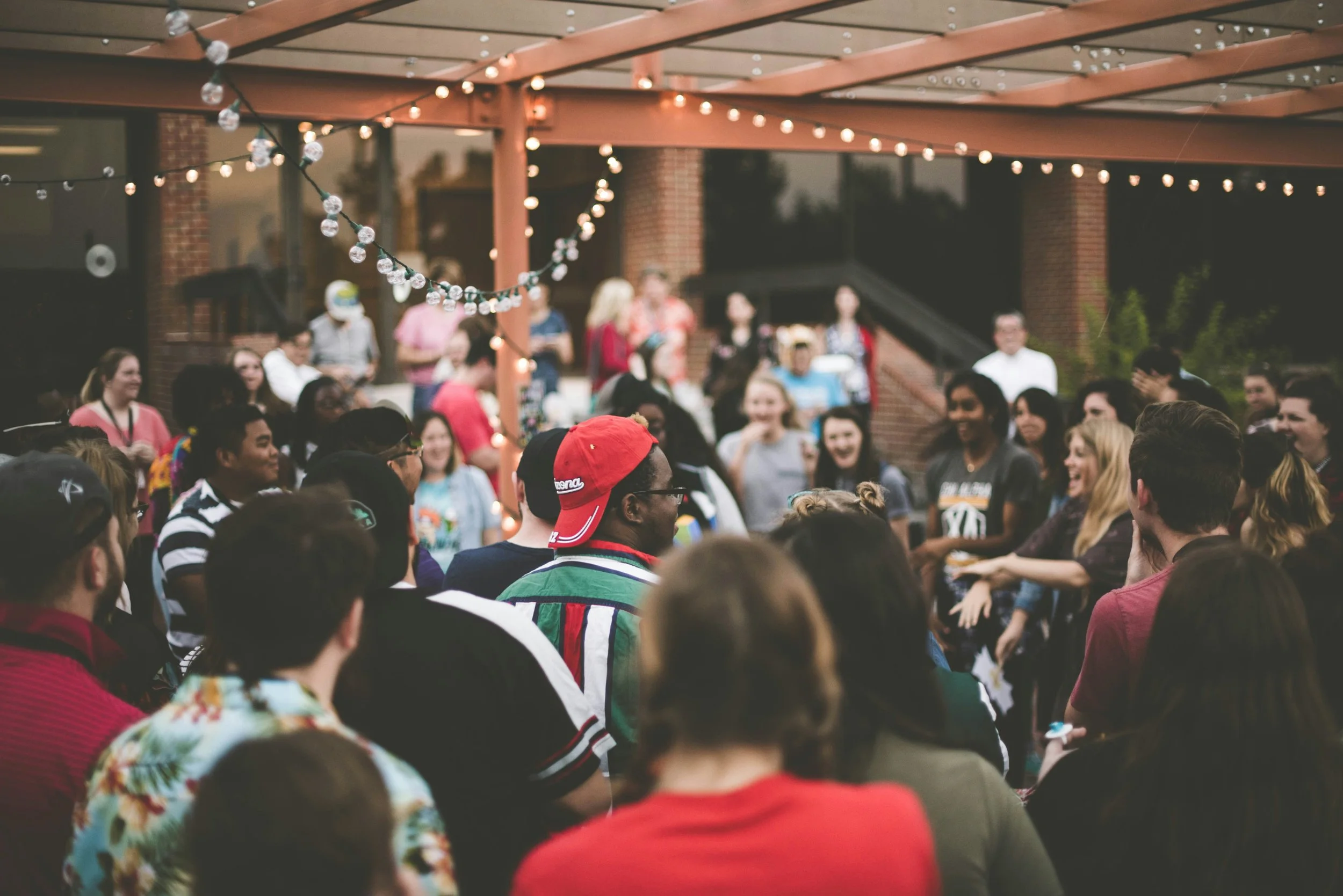 A group of people gathered outdoors under a wooden pergola decorated with string lights, with a brick building in the background. The scene appears to be a social event or celebration, with people smiling and engaging in conversations.