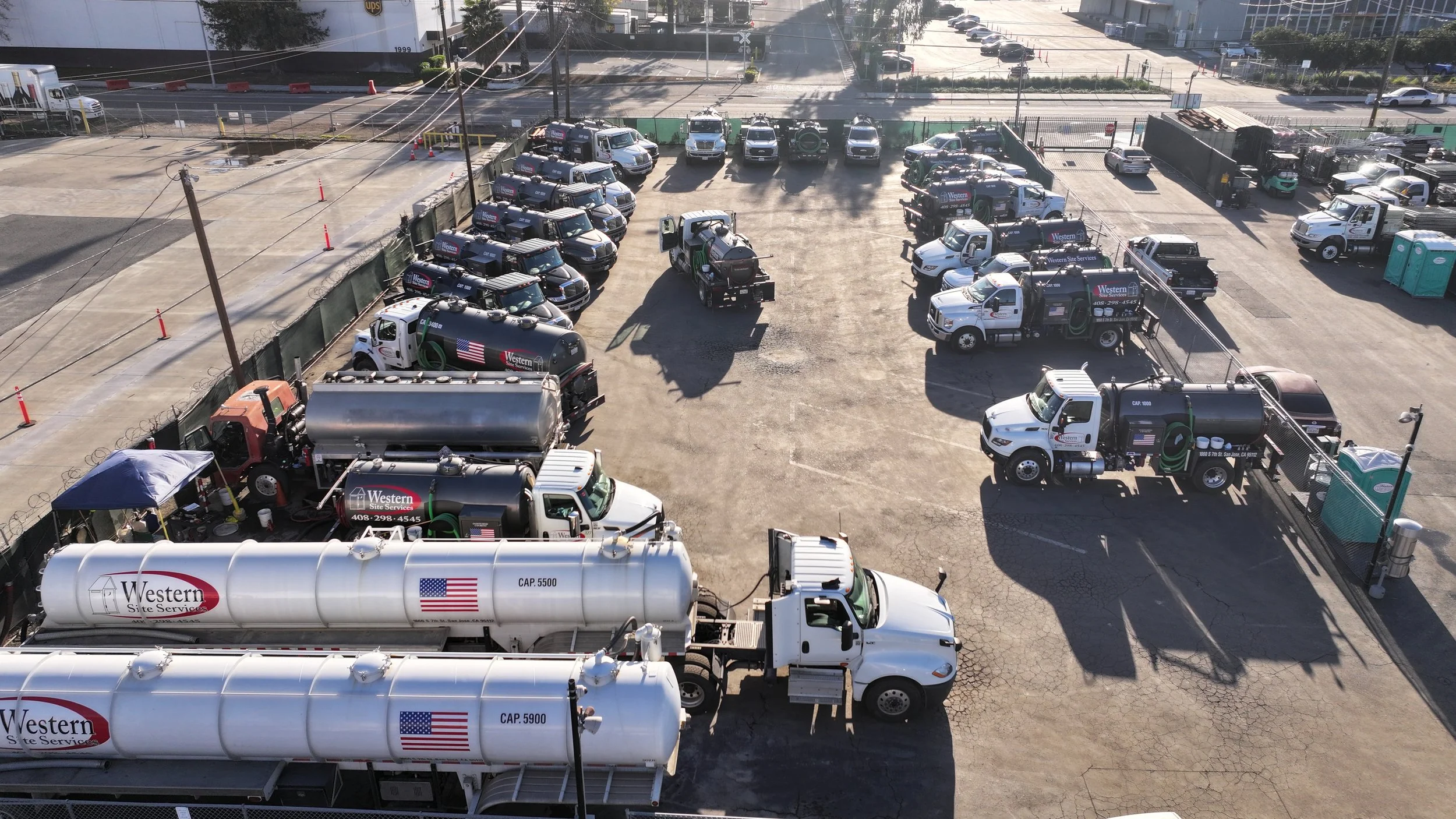 An aerial view of a parking lot with multiple service trucks and equipment stationed, some with Western Site Services branding and American flags, with a few portable toilets on right side and a street in the background.