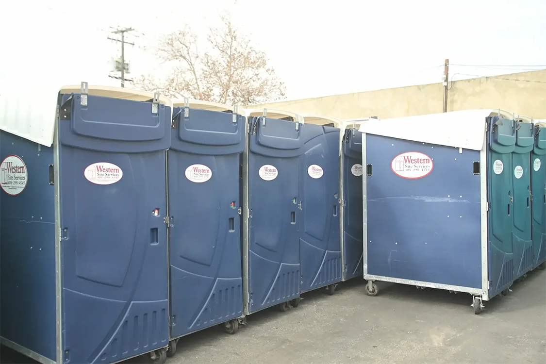 A row of high rise porta potties with company logo "Western Site Services" on the front, positioned on a paved outdoor area, with a beige wall and utility poles in the background.