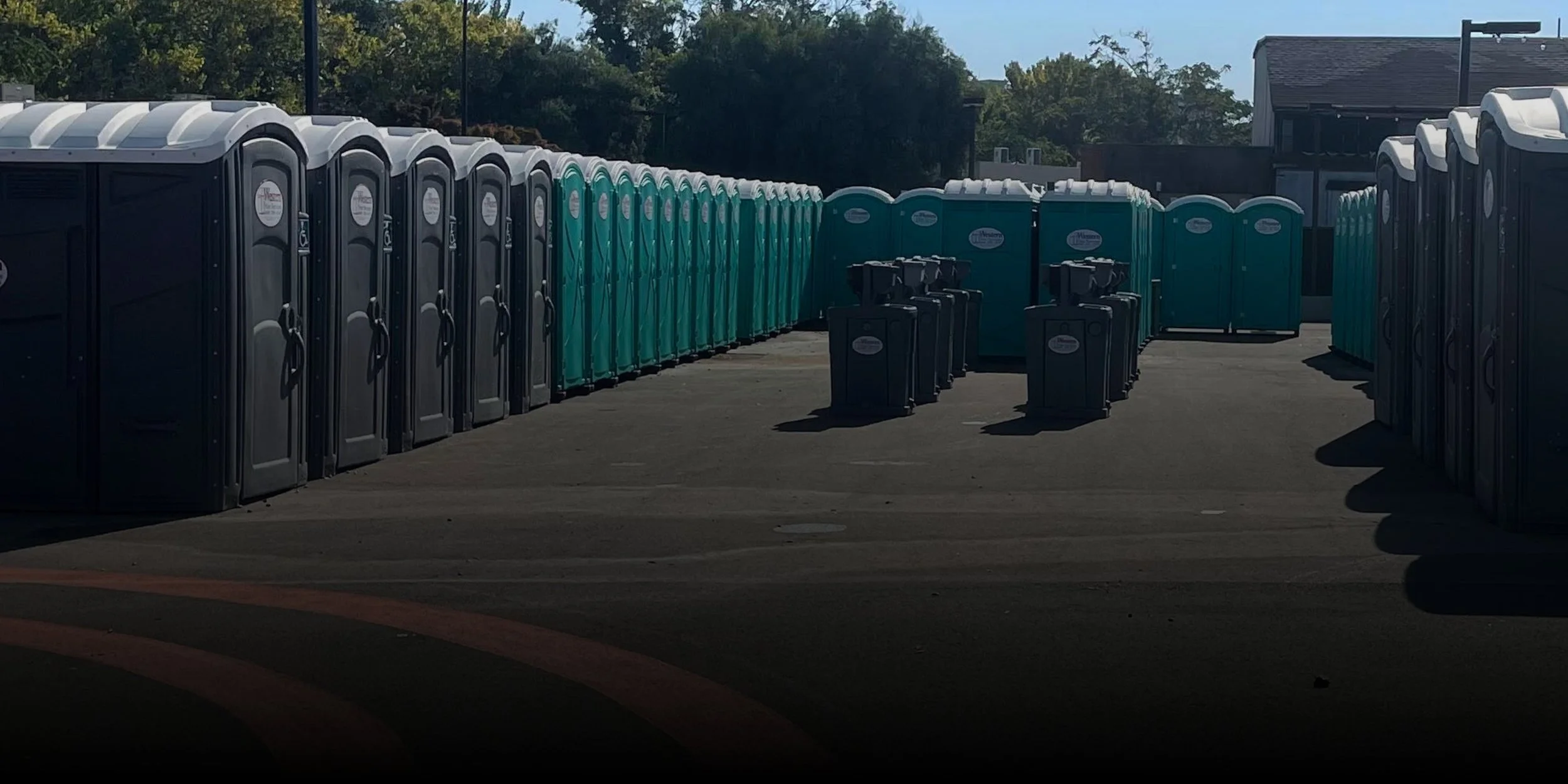 A row of portable toilets in black and teal colors, arranged in an outdoor area with a building and trees in the background.