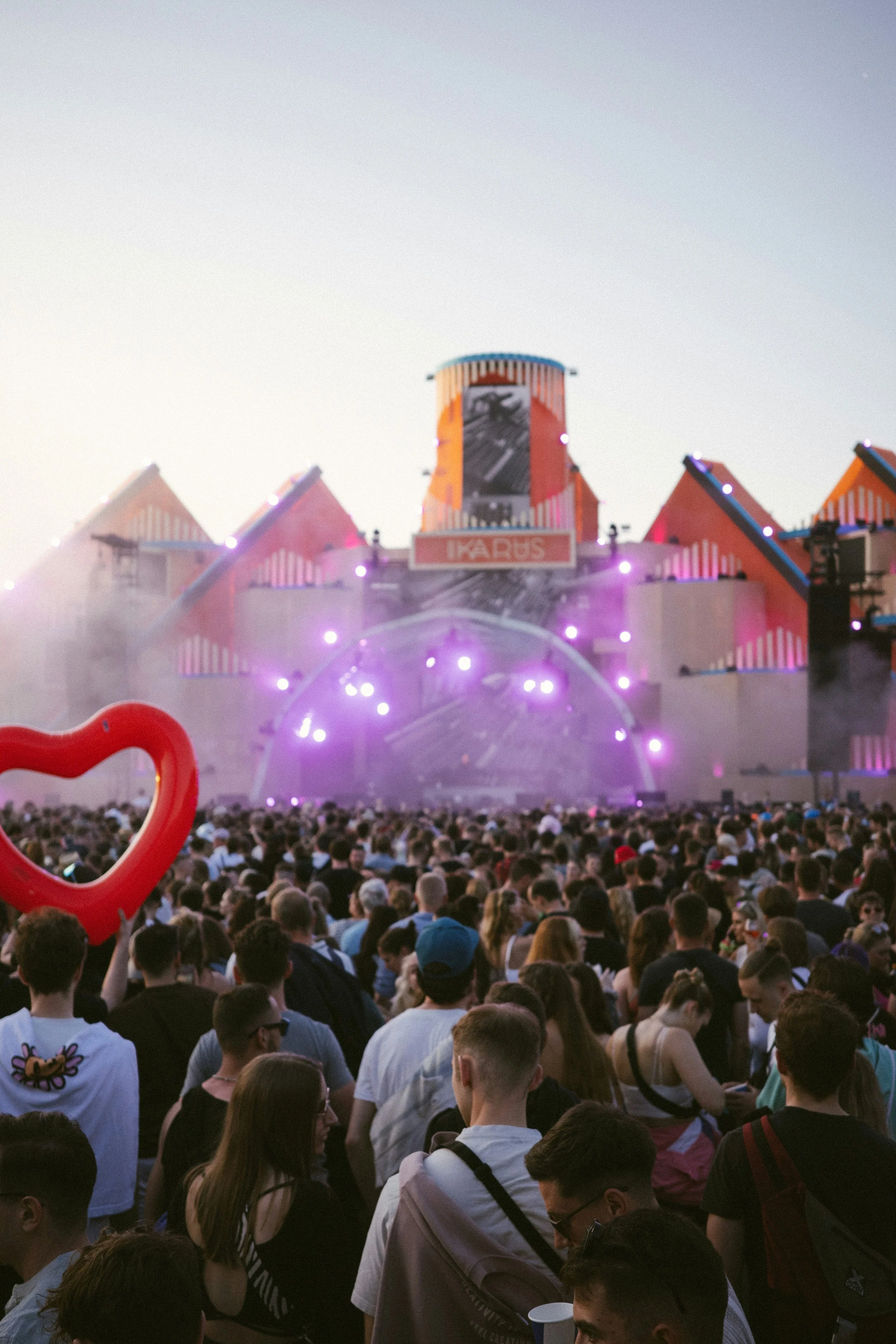 A large crowd of people attending an outdoor music festival with a stage in the background featuring pink, orange, and blue design elements and a sign that says "EDC RUS". Fog or smoke is visible at the front of the stage, and a pink heart-shaped balloon is held up in the crowd.