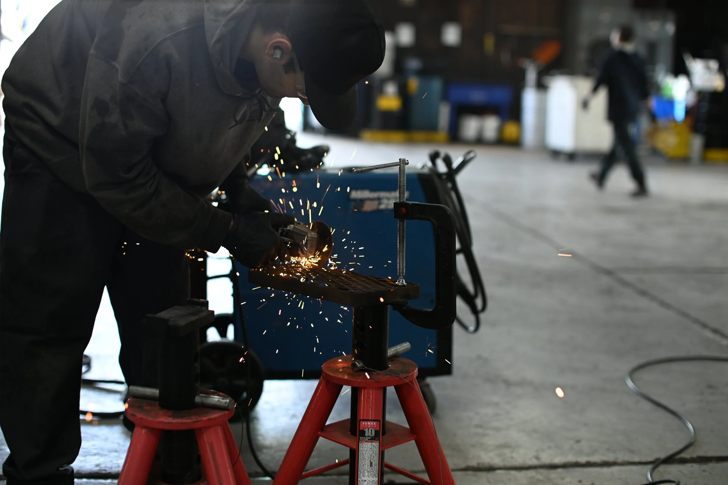A person is welding metal on a workbench in a workshop, with sparks flying. They are wearing protective gear, including a helmet and gloves. There are stools and other workshop equipment visible in the background.