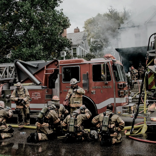 Firefighters in gear working in front of a red fire truck with smoke rising from a building