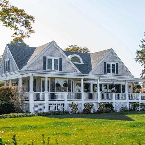 A large, white, two-story house with a wrap-around porch, multiple gabled roofs, and large windows, surrounded by a green lawn and some trees in the background.