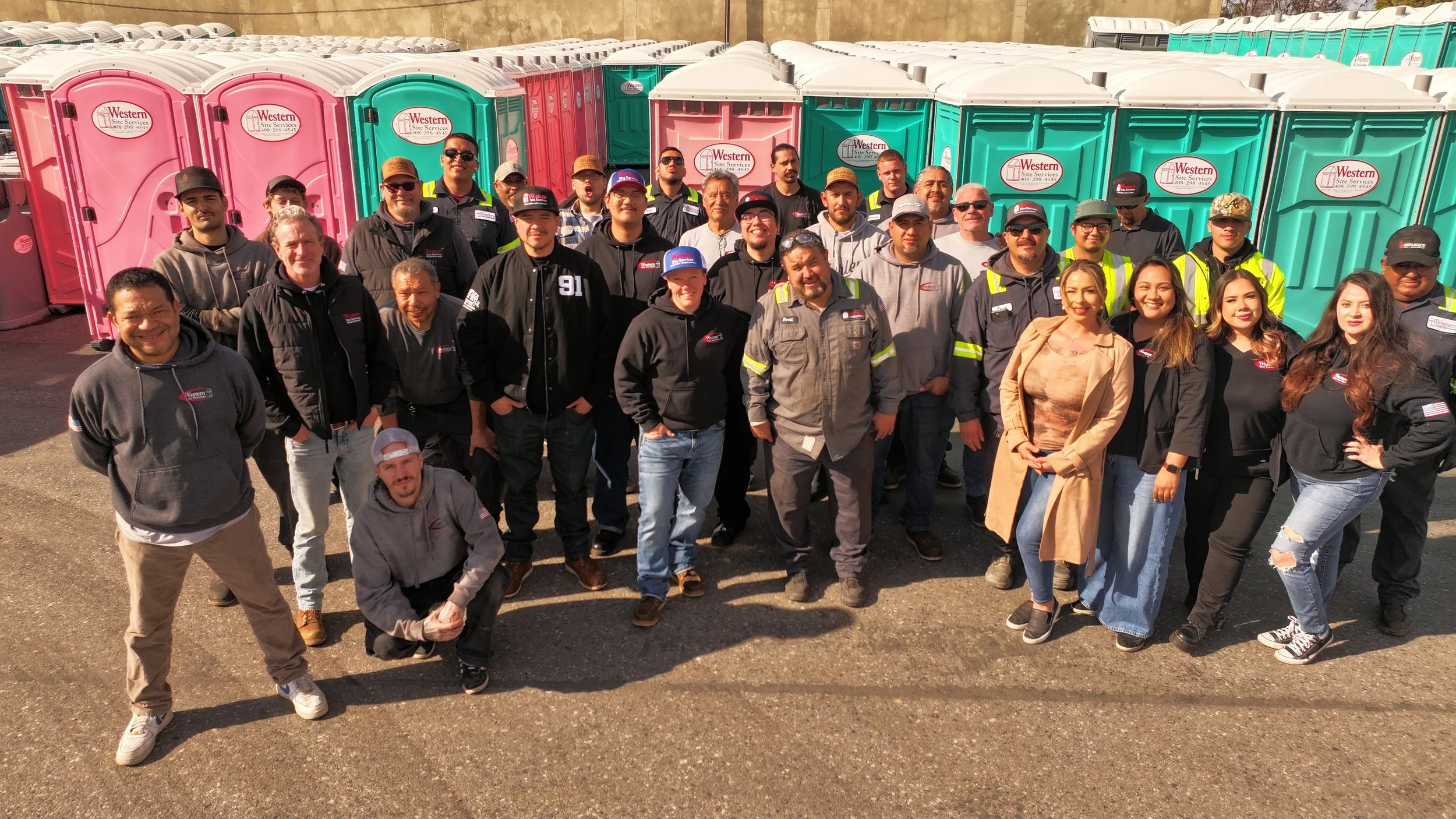 Group of people standing outdoors in front of colorful portable toilets, smiling at the camera.
