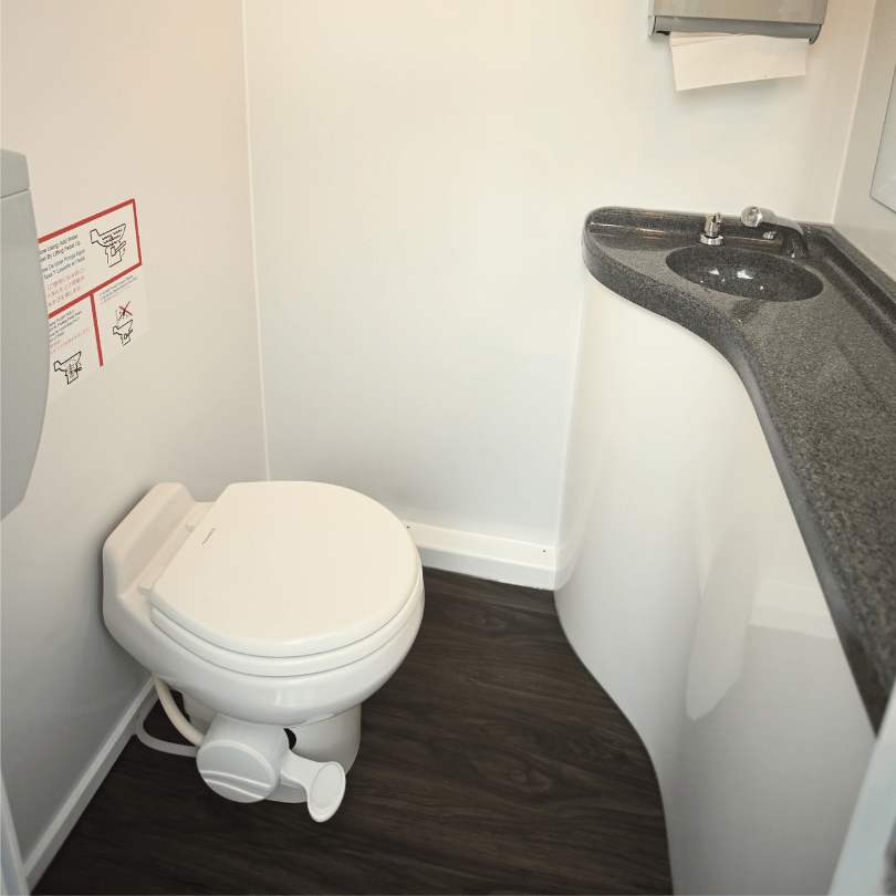 Small ADA portable bathroom with a white toilet and a black countertop with a sink, white walls, and a wood-look floor.