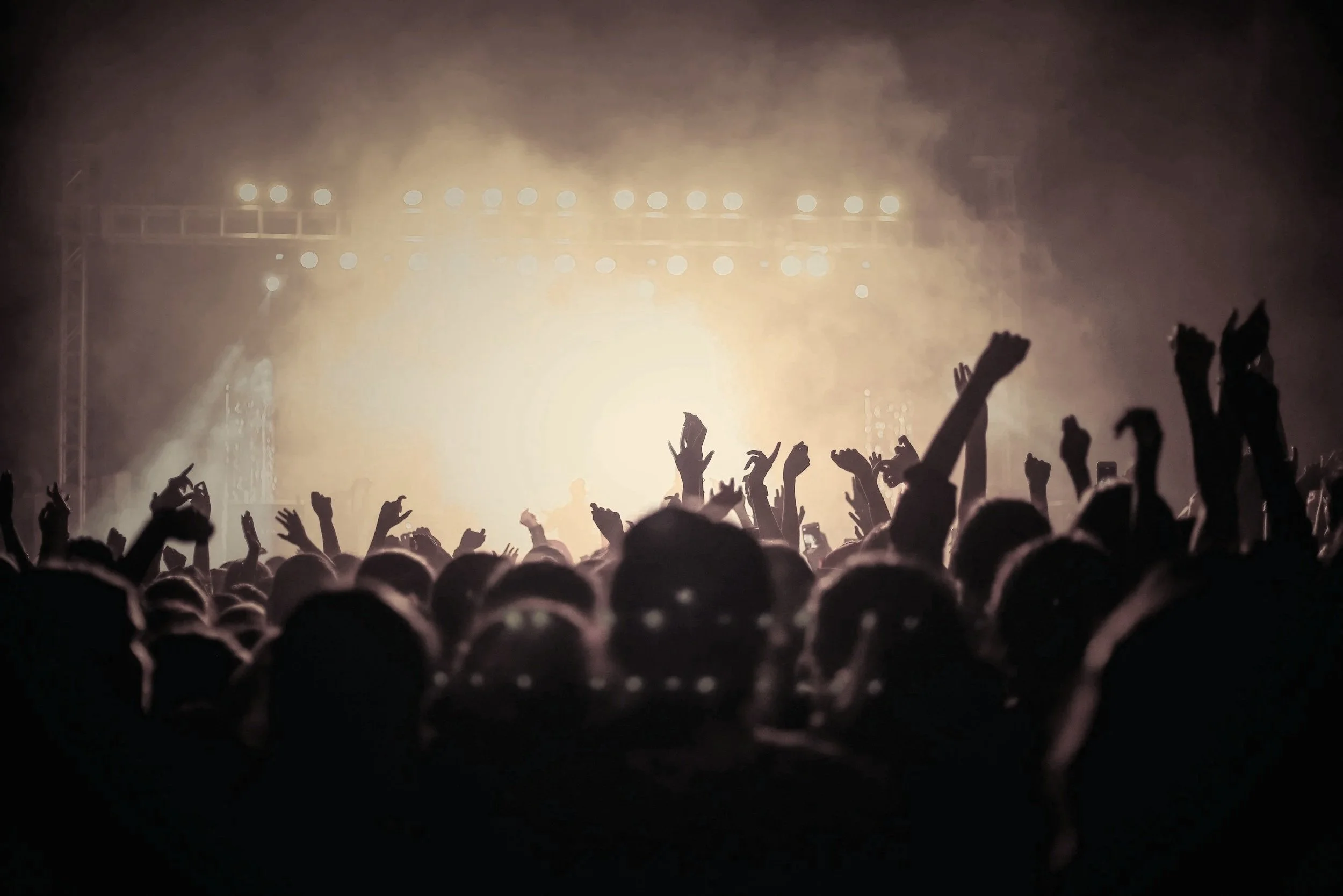 Crowd of concert-goers with hands raised, facing a brightly lit stage with fog and numerous lights in dark atmosphere.