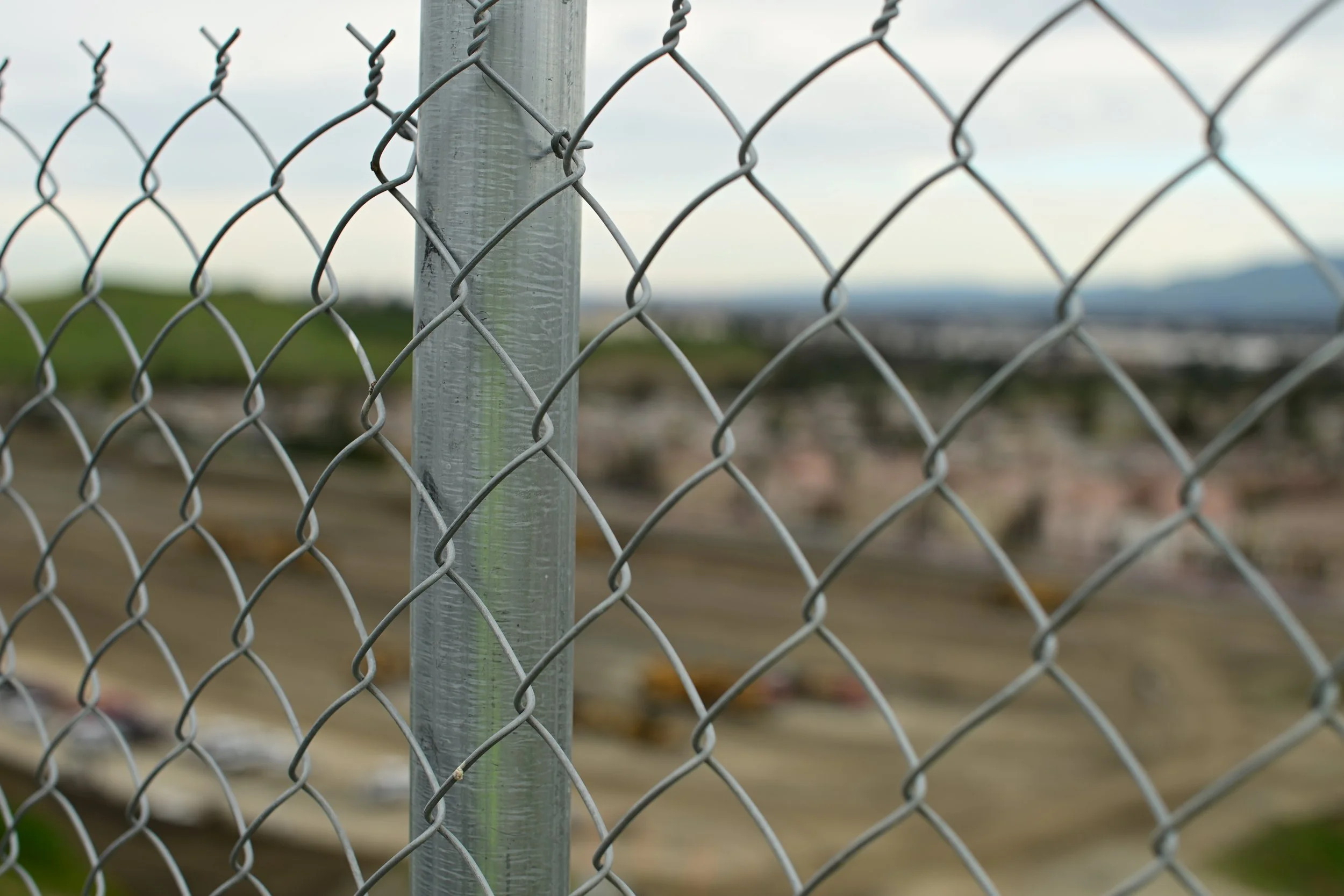 Close-up of a metal chain-link fence with a metal post in the foreground. The background is blurred, showing a landscape with green hills and mountains under an overcast sky.