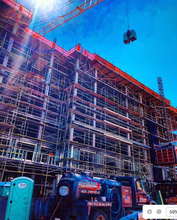 Construction site with a multi-story building under construction, scaffolding surrounding it, and a crane lifting materials, with a blue sky in the background.