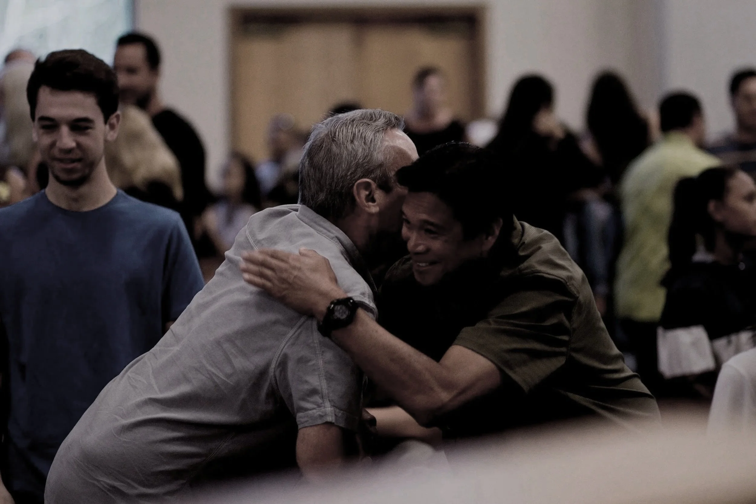 Two men hugging each other warmly while others stand around in a crowded room, smiling and socializing.