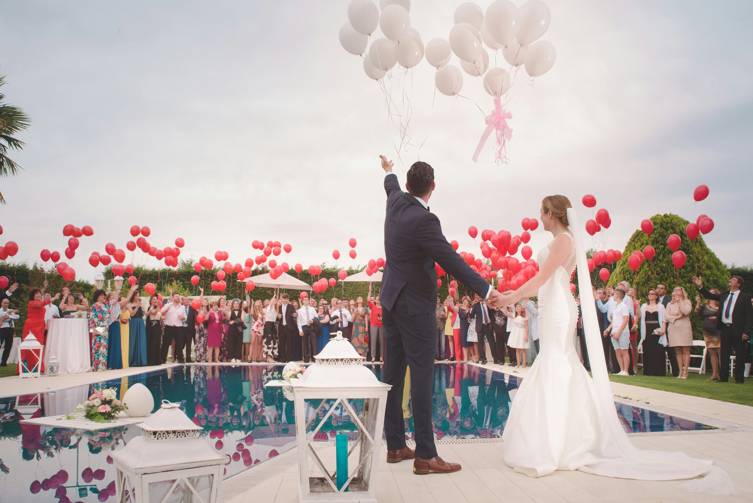 A wedding ceremony outdoors with a bride and groom holding hands, surrounded by many guests. The groom is tossing white balloons into the air, and there are red balloons in the background. The scene is festive with decorated tables and a clear sky.