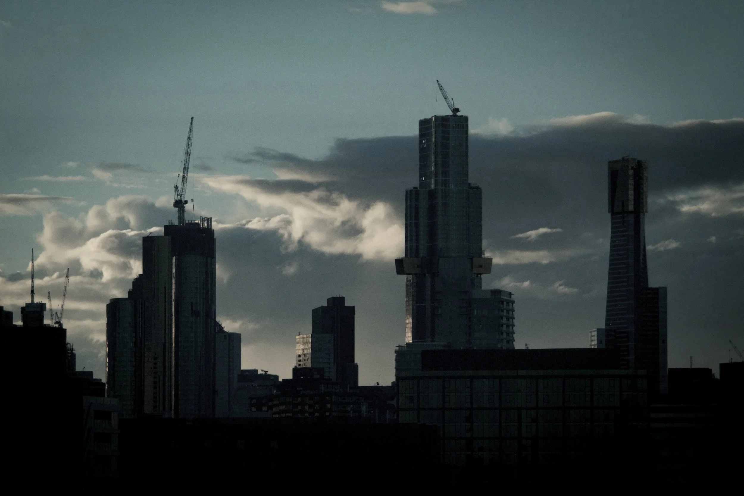 A city skyline during the evening or late afternoon with several tall skyscrapers under construction, some with cranes on top, set against a cloudy sky.