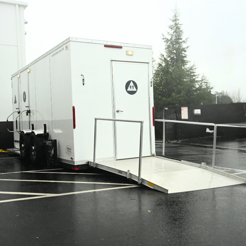 A white portable restroom trailer with accessibility features, including a ramp and handrails, parked in an outdoor lot on a rainy day.