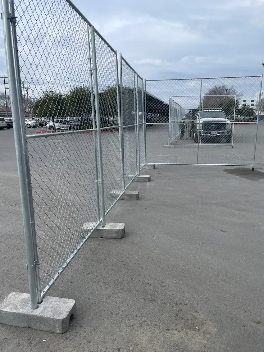 A chain-link fence section with a gate in a parking lot, few cars visible, overcast sky.