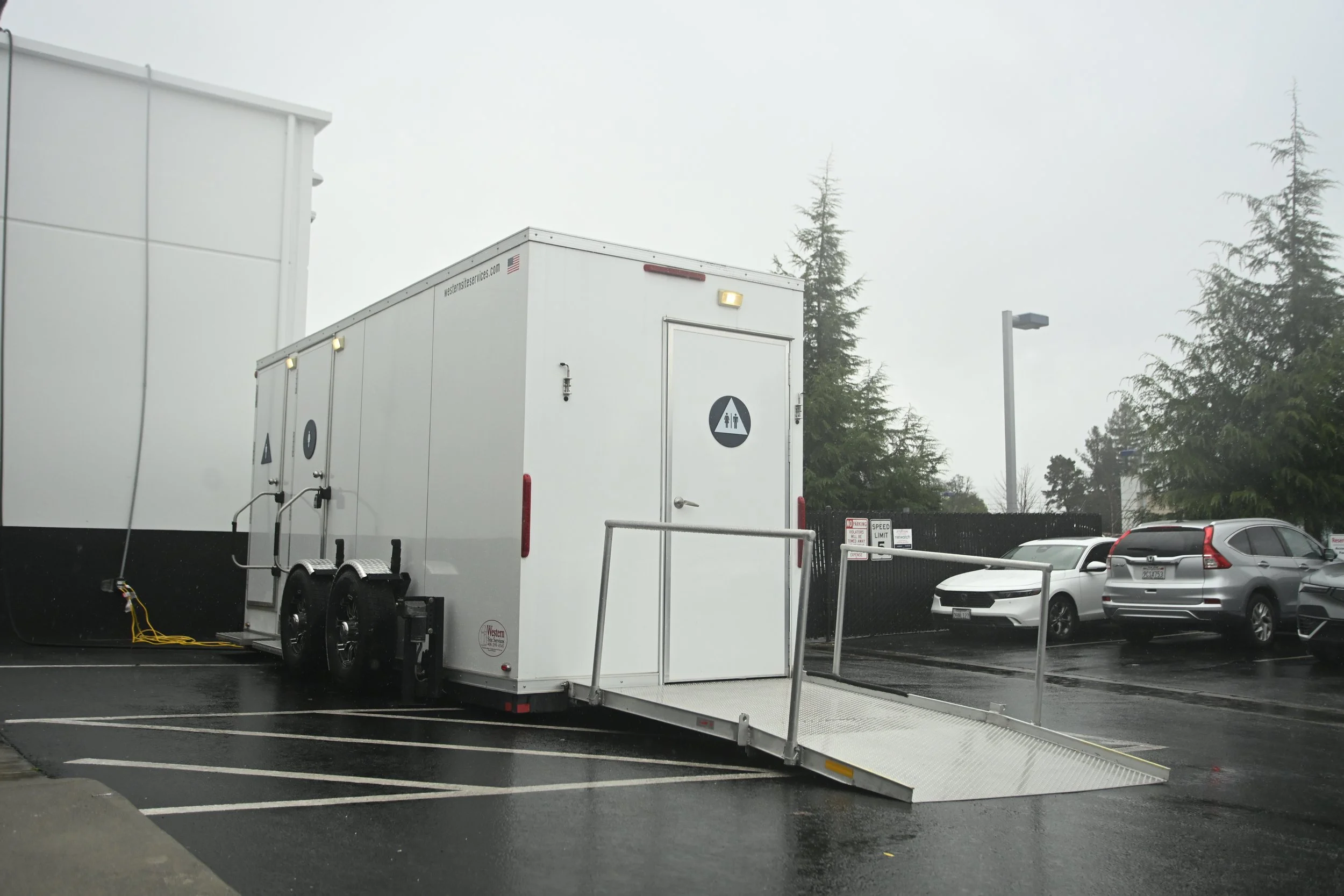 A white mobile restroom trailer in a parking lot, with a wheelchair ramp extended, surrounded by cars and tall trees, on an overcast day.