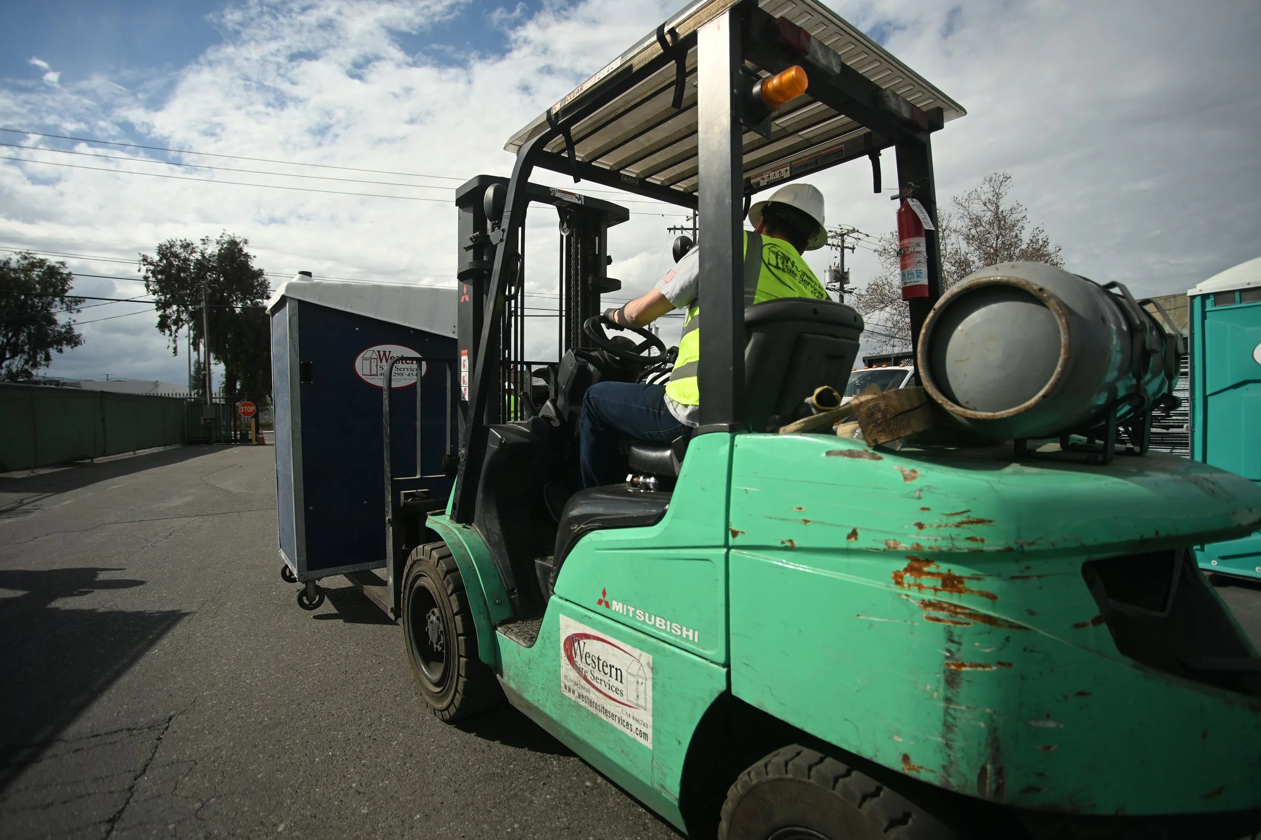 A person wearing a white helmet and fluorescent safety vest operating a green Mitsubishi forklift with a fire extinguisher mounted nearby, in an outdoor industrial area under a cloudy sky.