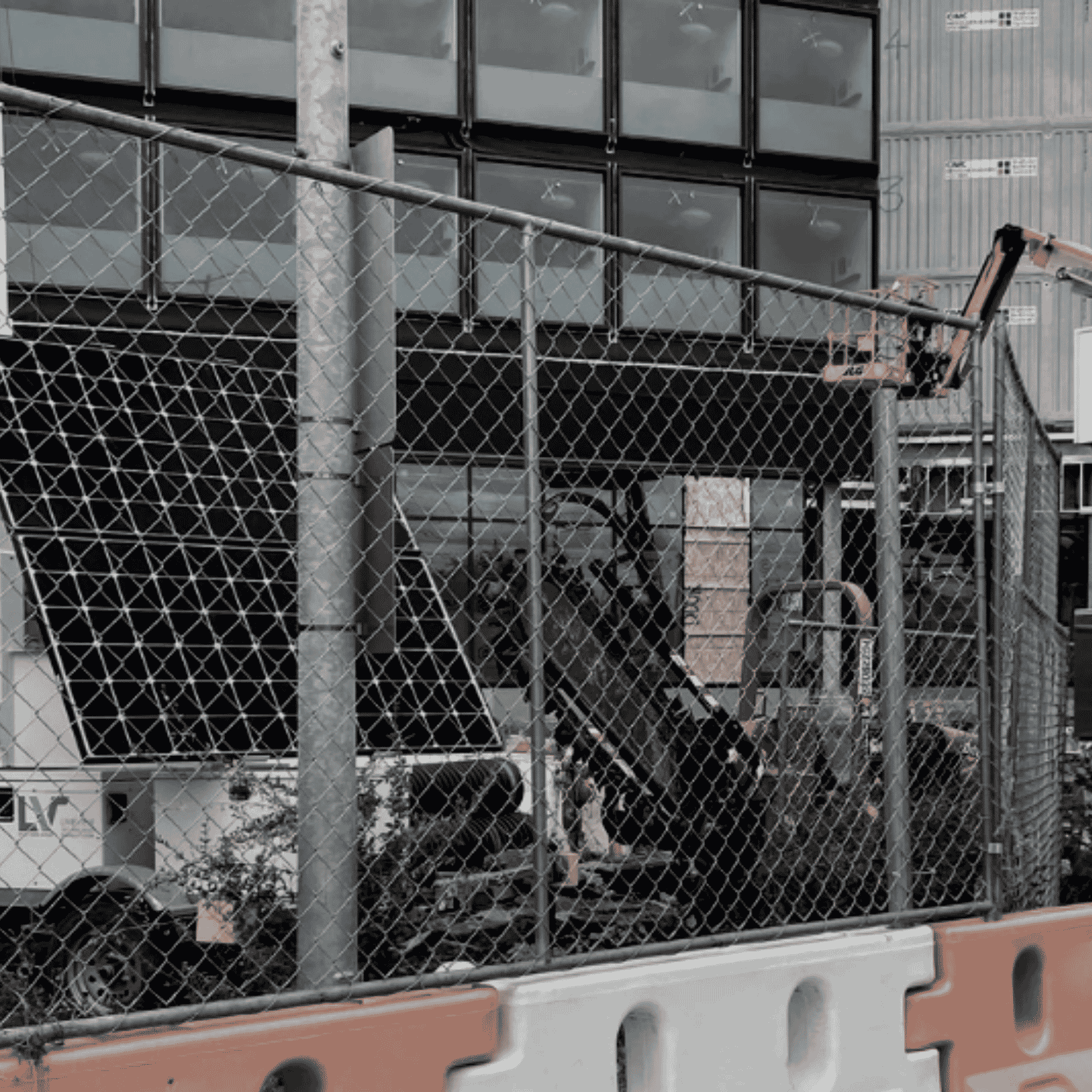 A construction site with a building under construction, visible behind a chain-link fence. Construction equipment and materials are present, including a hydraulic lift and boxes.