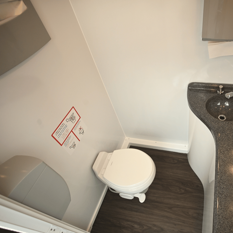 Inside of a small portable restroom trailer with a white toilet, a gray countertop with a black sink, and a partially visible paper towel dispenser on the wall.