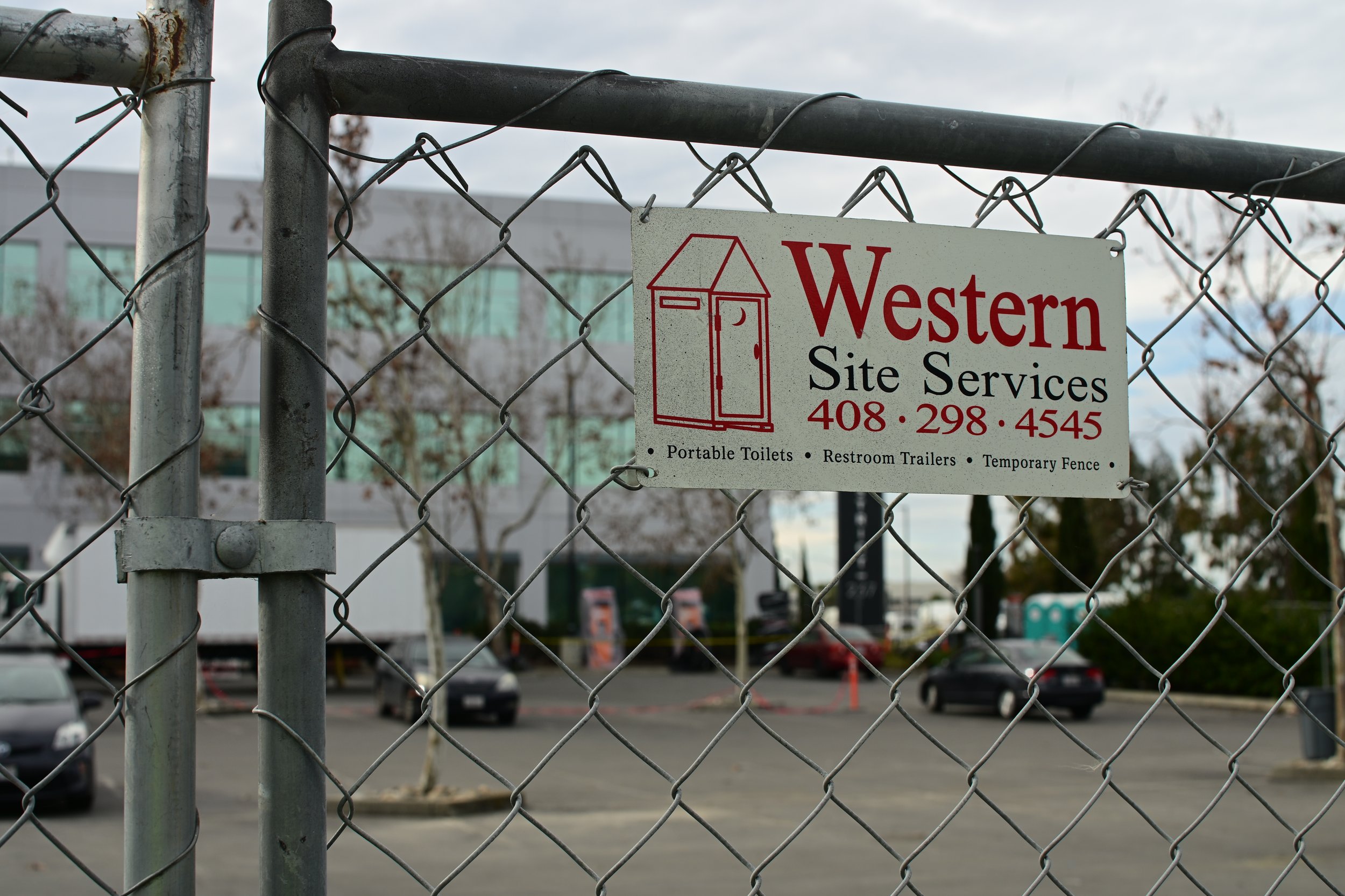 A metal chain-link fence with a sign for Western Site Services, displaying a phone number and icons for portable toilets, restroom trailers, and temporary fencing. In the background, a parking lot with a few cars and a building with many windows are visible.