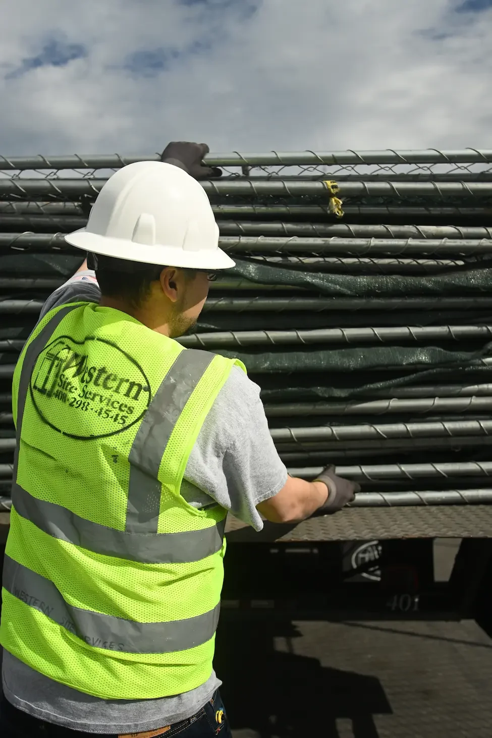 A construction worker wearing a white hard hat, a yellow high-visibility vest, and gloves is handling a bundle of black electrical poles or pipes on a flatbed trailer, under a partly cloudy sky.