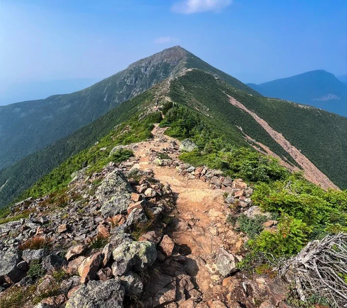 A mountain trail leading to a peak, surrounded by green shrubs and rocks under a blue sky.