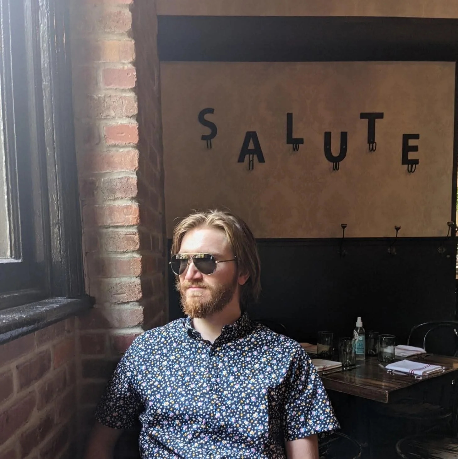 A man with shoulder-length hair, sunglasses, and a beard sitting in a restaurant near a brick wall with a large window. The background has a sign with the word 'SALUTE' in black letters.