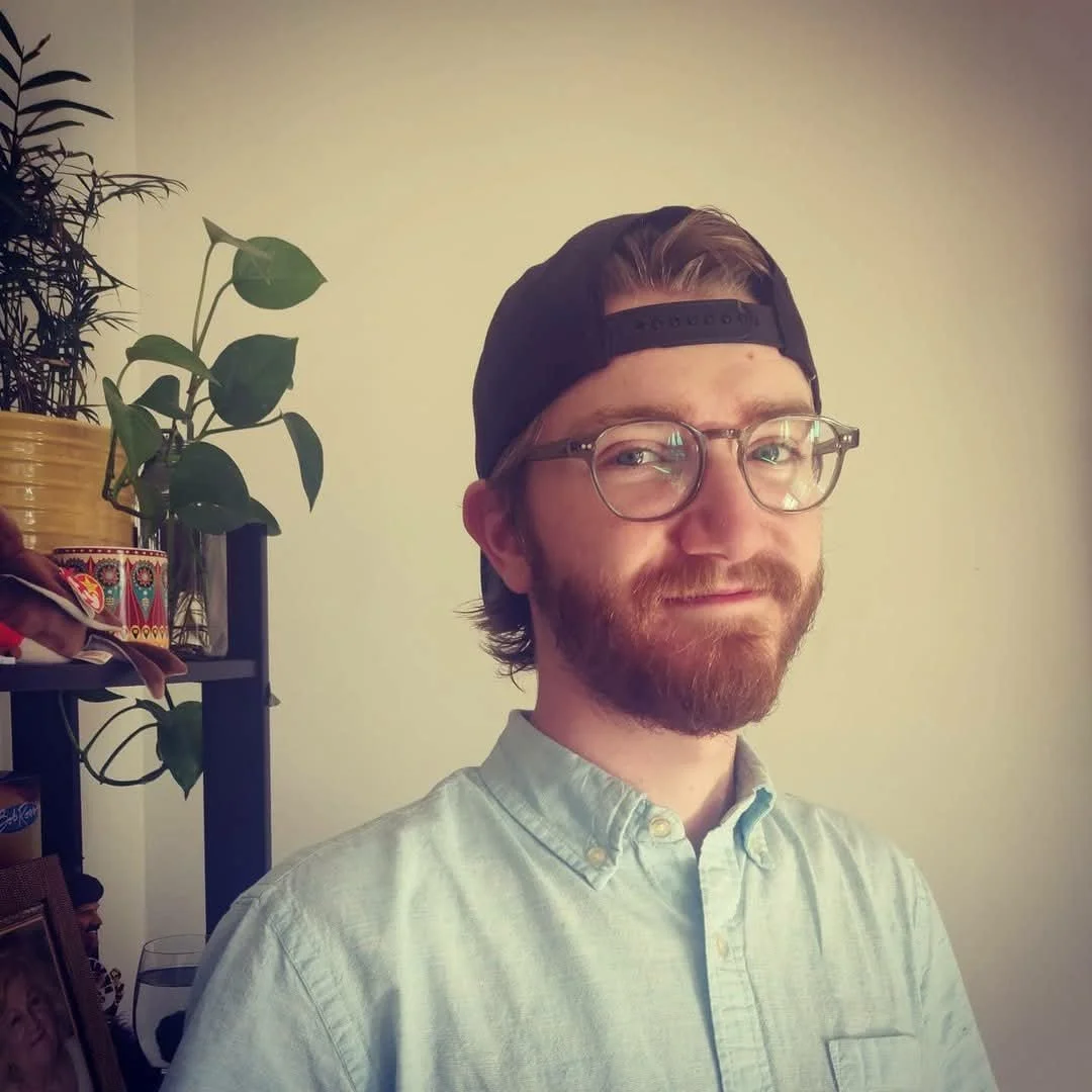 A man with glasses, a beard, and a backwards baseball cap, smiling indoors with a plant and a shelf in the background.