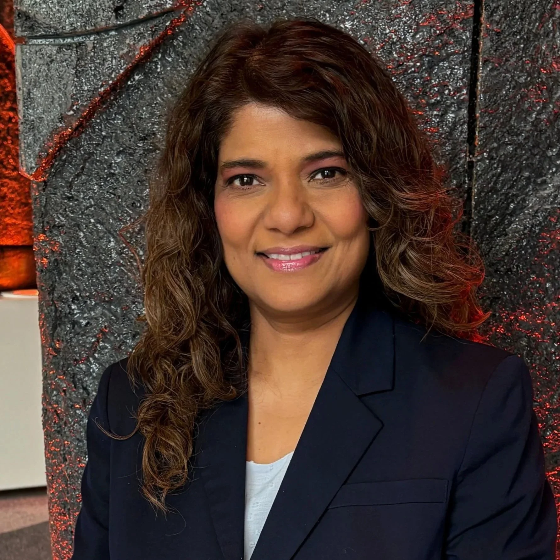 A woman with curly brown hair, wearing a black blazer and white top, smiling in front of a textured, 3D gray wall background with red lighting.