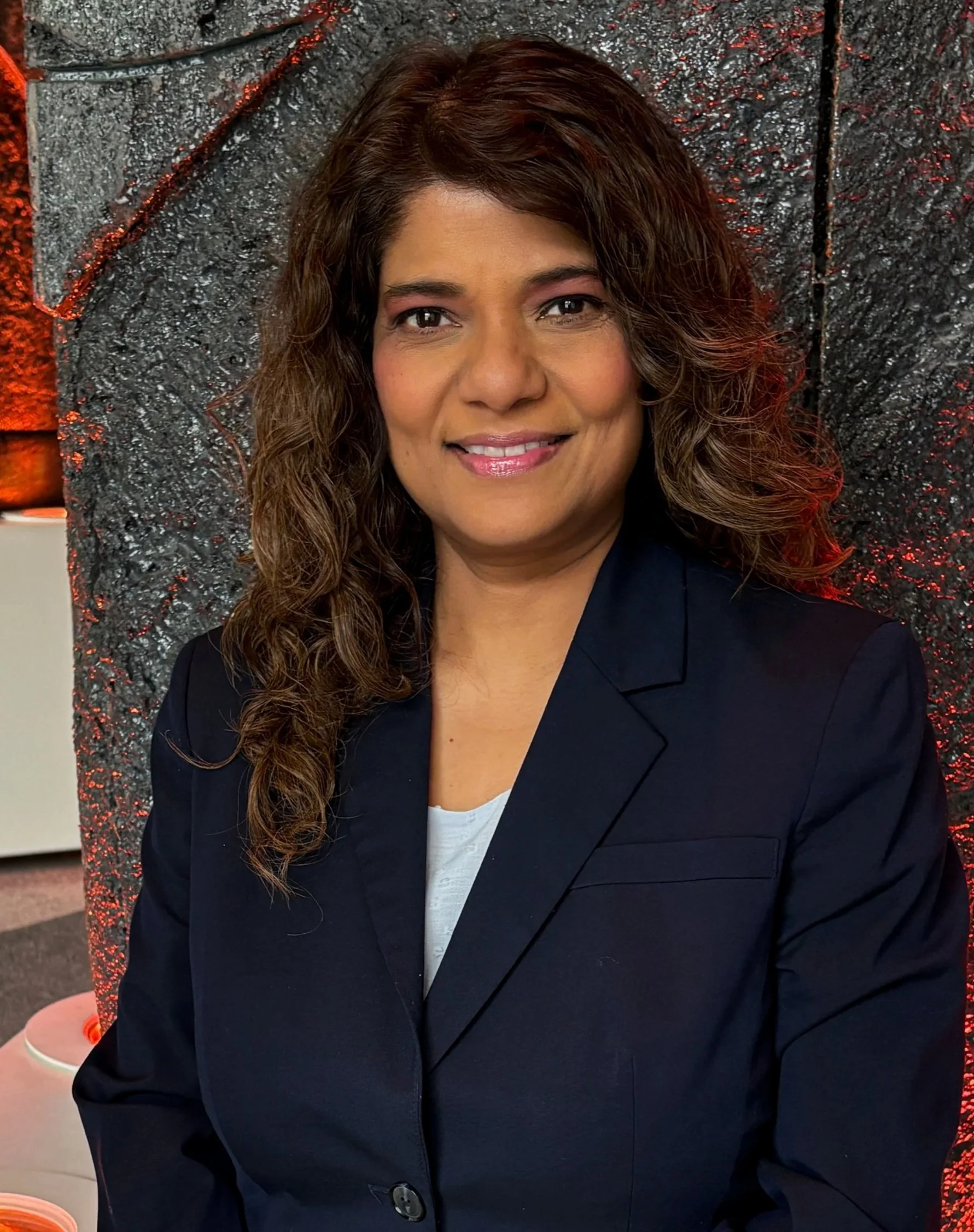 A woman with long wavy brown hair wearing a dark blazer and light-colored top, smiling and posing indoors against a textured black and gray background with red lighting accents.