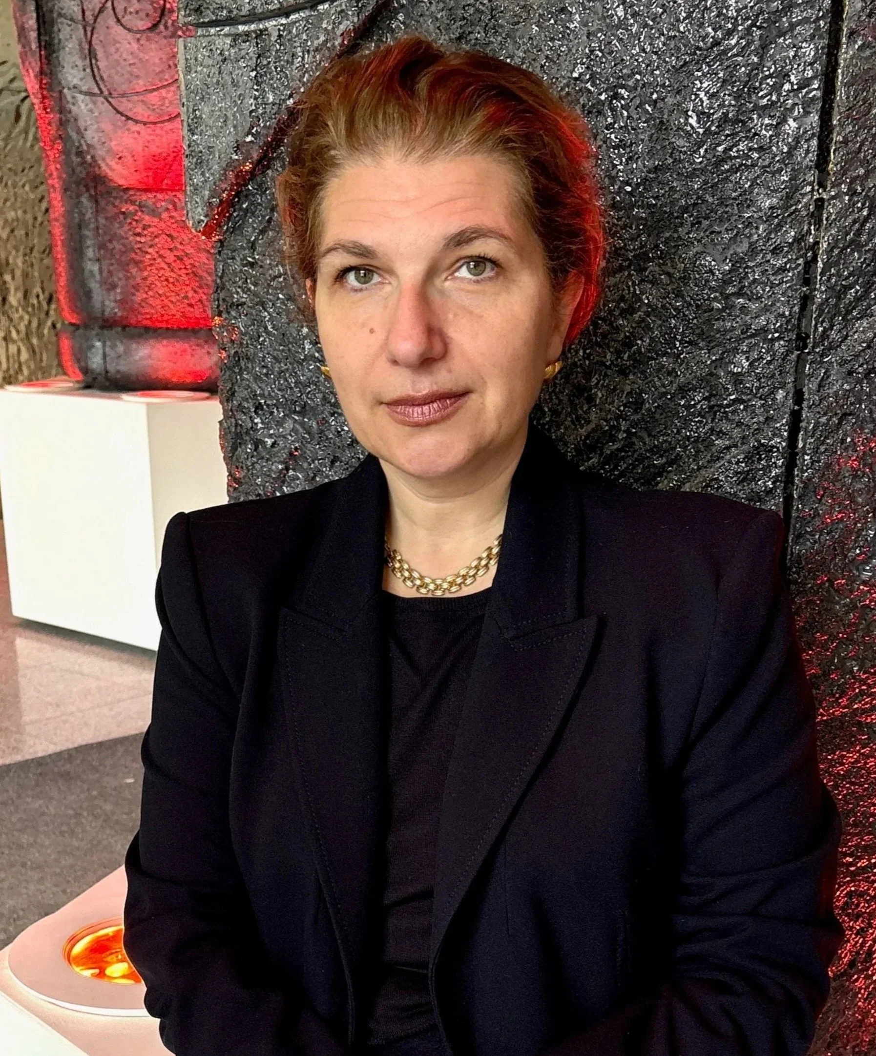 A woman with short, reddish hair wearing a black blazer and gold chain necklace, sitting against a textured black wall.