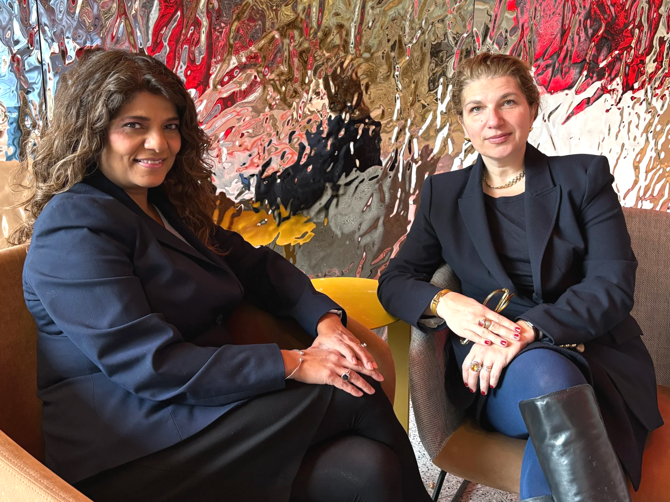 Two women seated on chairs, smiling at the camera, with an abstract colorful wall behind them.