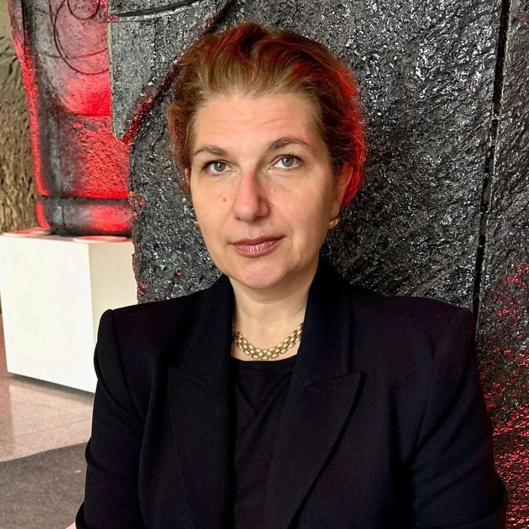 A woman with short reddish hair, wearing a black blazer and a gold chain necklace, seated against a black textured wall.