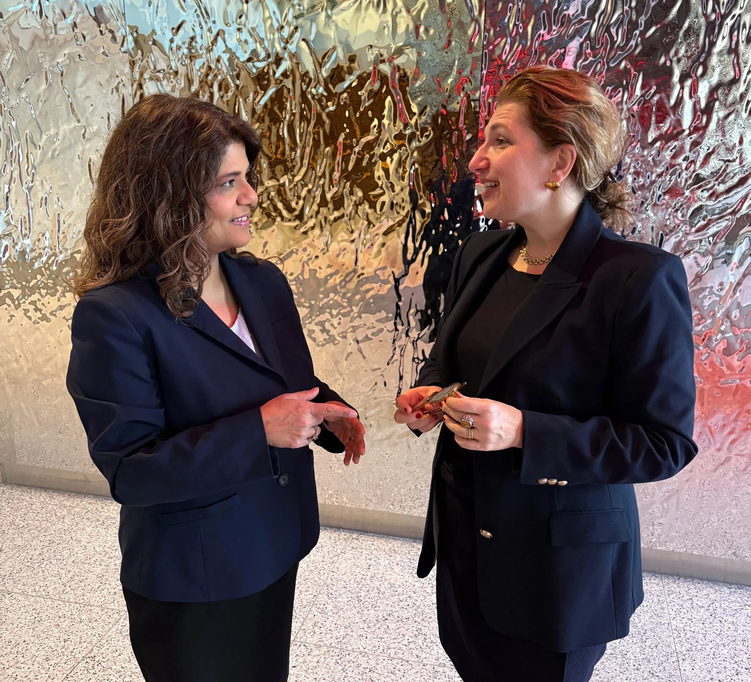 Two women in formal business suits having a conversation and smiling at each other, standing in front of an abstract, metallic wall art.