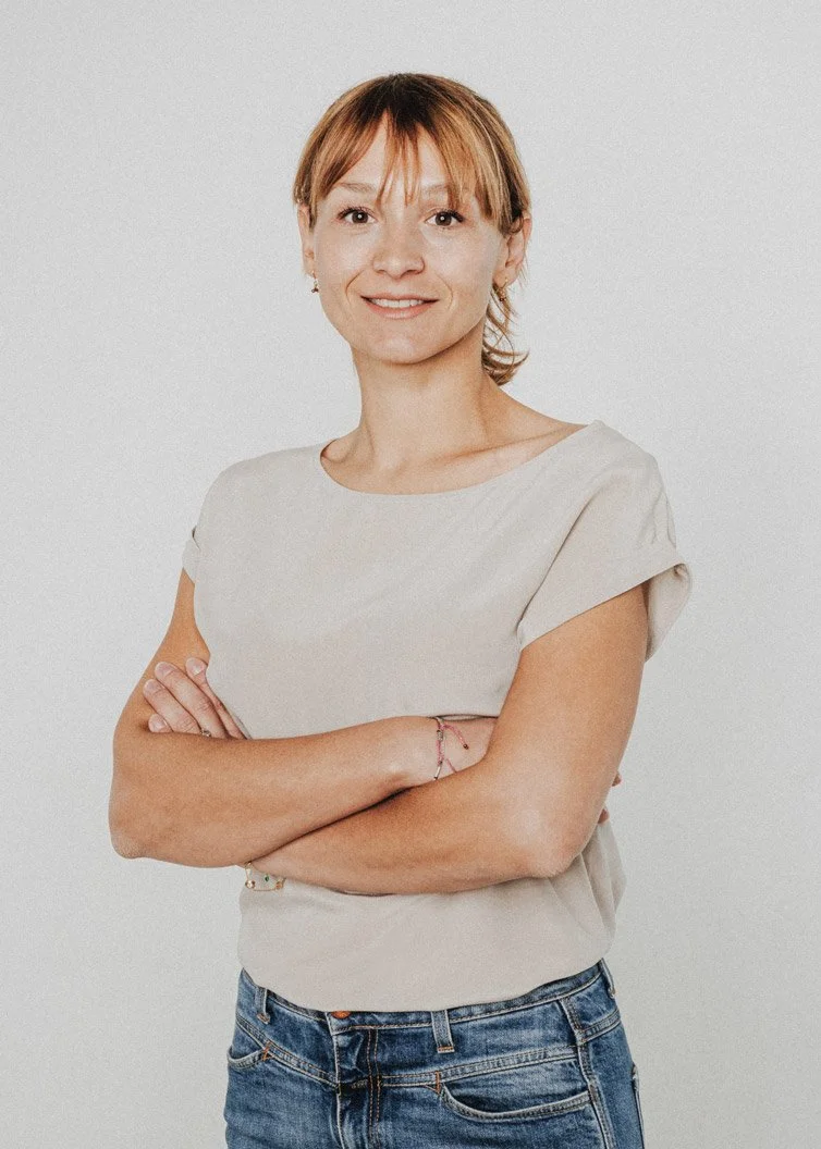 Eine Frau mit braunen Haaren trägt ein beiges T-Shirt und Jeans, steht vor einem weißen Hintergrund, lächelt leicht und hat die Arme verschränkt.
