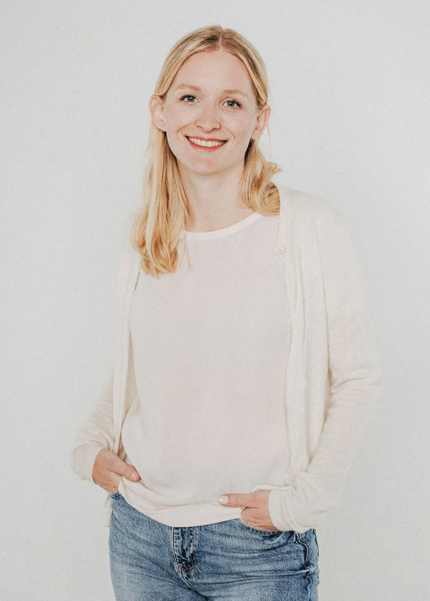 Eine junge Frau mit blonden Haaren, Lächeln, weißem T-Shirt, heller Jacke und blauen Jeans vor weißem Hintergrund.