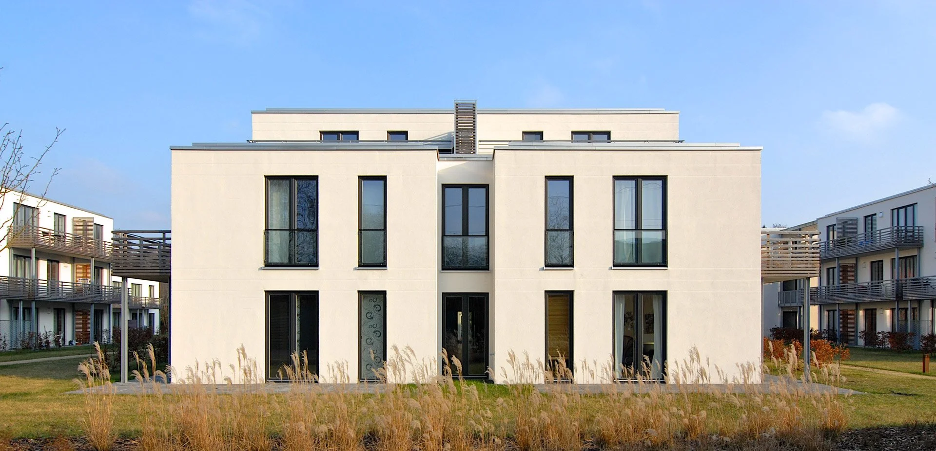 Moderne weiße Wohnanlage mit mehreren Stockwerken, große Fenster und Balkonaufbauten, umgeben von Grünflächen, bei sonnigem Himmel.